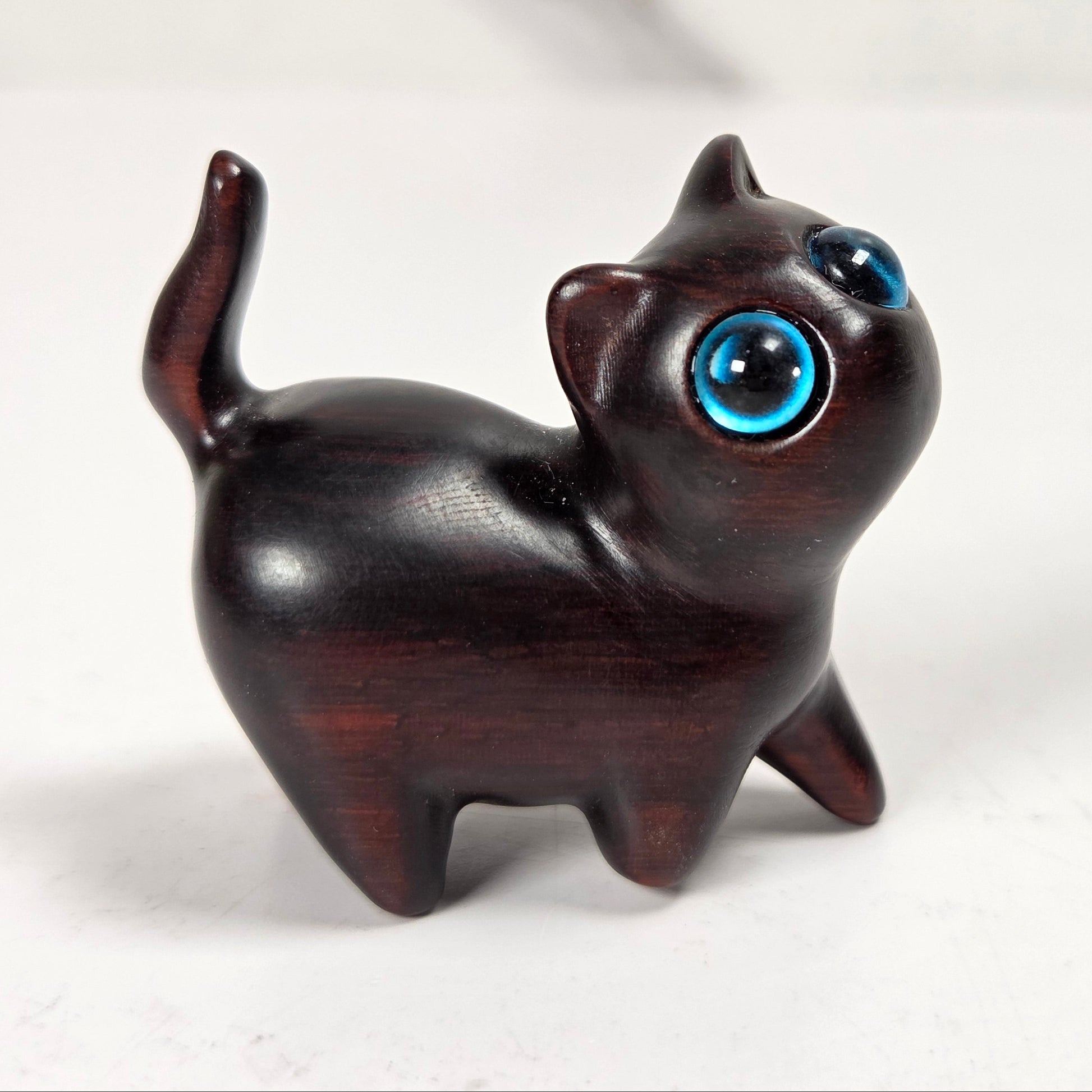 A small carved wooden cat figurine with a glossy dark brown finish and large, expressive blue eyes, standing on a light surface against a soft white background.