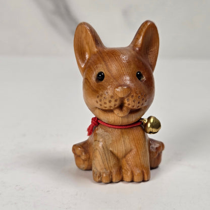 A small carved wooden dog figurine with a red collar and a small gold bell, photographed on a light neutral surface.