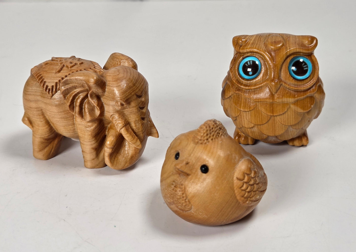 Three small carved wooden animal figurines on a white surface: an elephant, an owl with large blue eyes, and a bird, photographed against a light marble-look background.