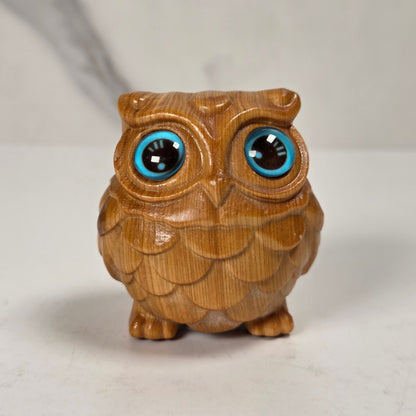 A small carved wooden owl figurine with large, glossy blue and black eyes, standing on a light surface against a neutral background.