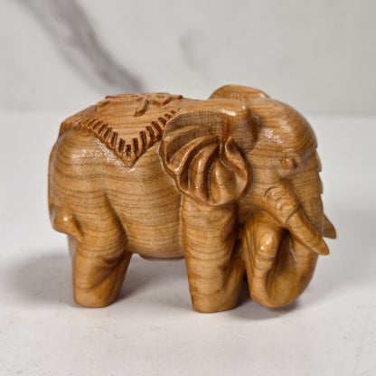 A small carved wooden elephant figurine with detailed trunk, tusks, and decorative back patterns, shown on a light marble-like surface.