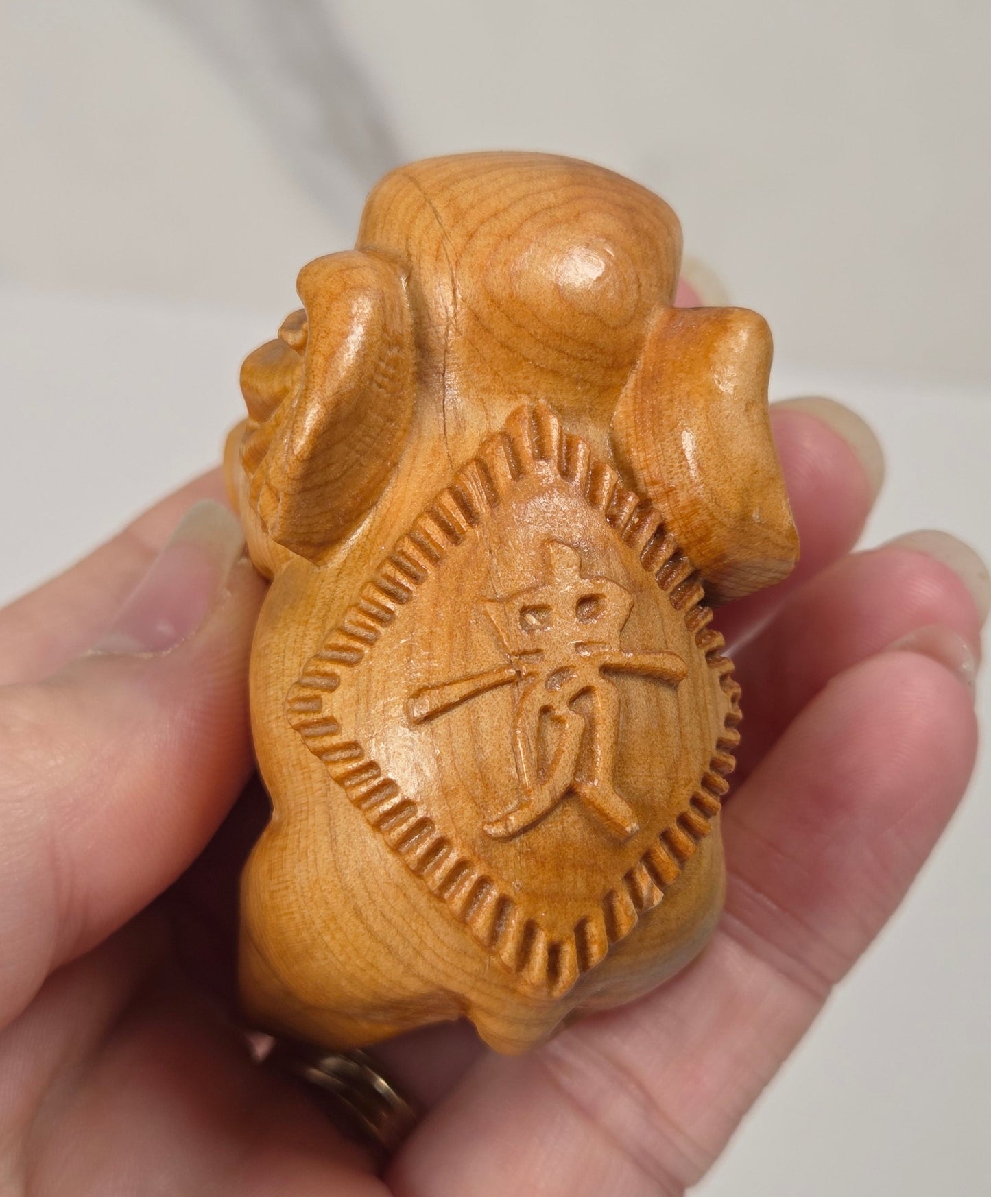 A hand holding a small carved wooden figurine with a golden-yellow finish and an engraved Chinese character on the front, set against a light marble-patterned background.