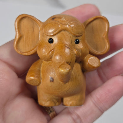 A small carved wooden elephant figurine with heart-shaped ears, held in a person's hand against a light gray background.
