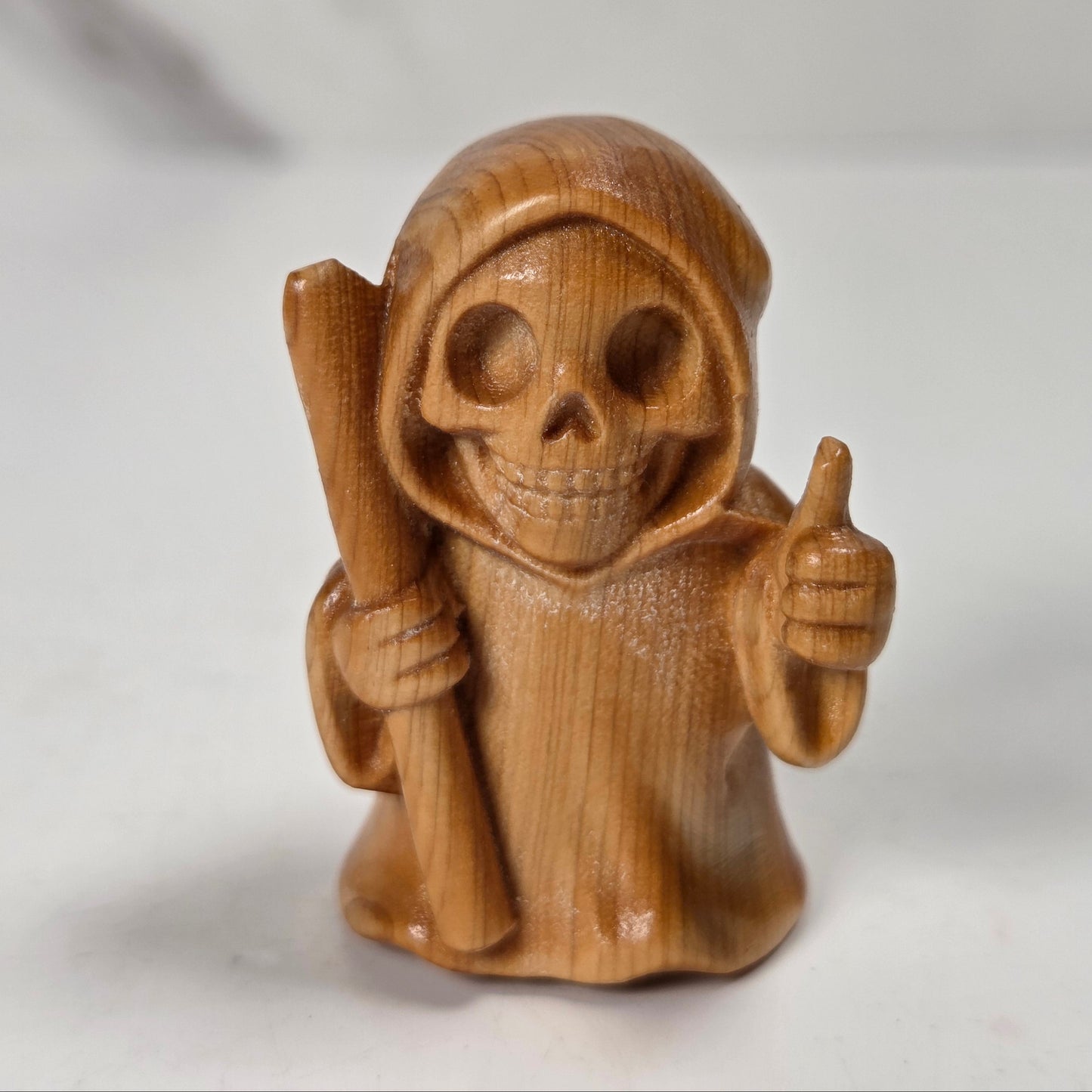 A small carved wooden grim reaper figurine with a skull face, holding a staff and giving a thumbs-up, shown on a light gray surface.