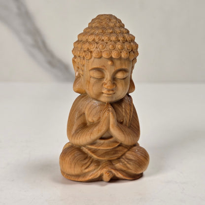 A small wooden Buddha figurine in a seated meditation pose, crafted from light brown wood with visible grain, set against a neutral light gray background.