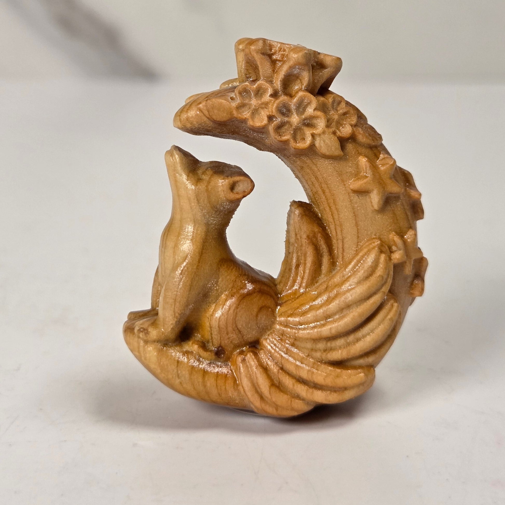 A small carved wooden figurine of a fox sitting on a crescent moon with floral details, photographed on a light gray surface with a blurred neutral background.