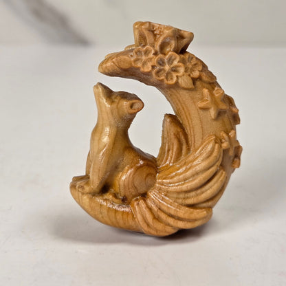 A small carved wooden figurine of a fox sitting on a crescent moon with floral details, photographed on a light gray surface with a blurred neutral background.