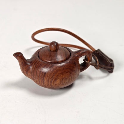 A small hand-carved wooden teapot with a dark brown wood grain finish and a matching leather strap, photographed on a plain off-white background.