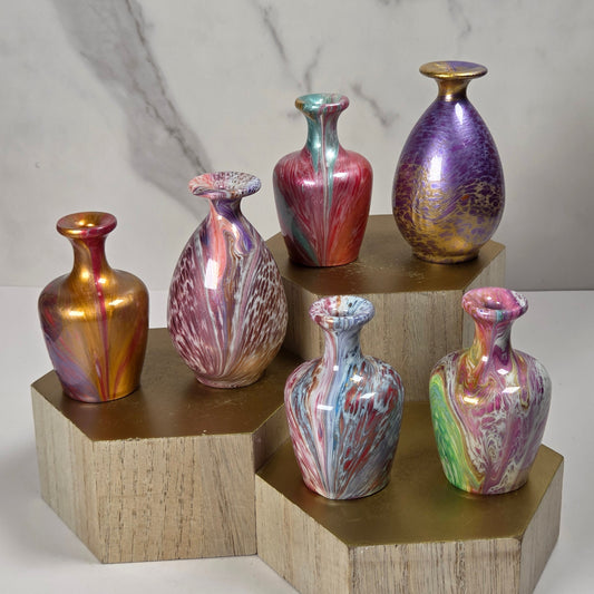 Seven iridescent glass vases in assorted colors and shapes, displayed on a tiered wooden riser against a light gray marble-patterned background.