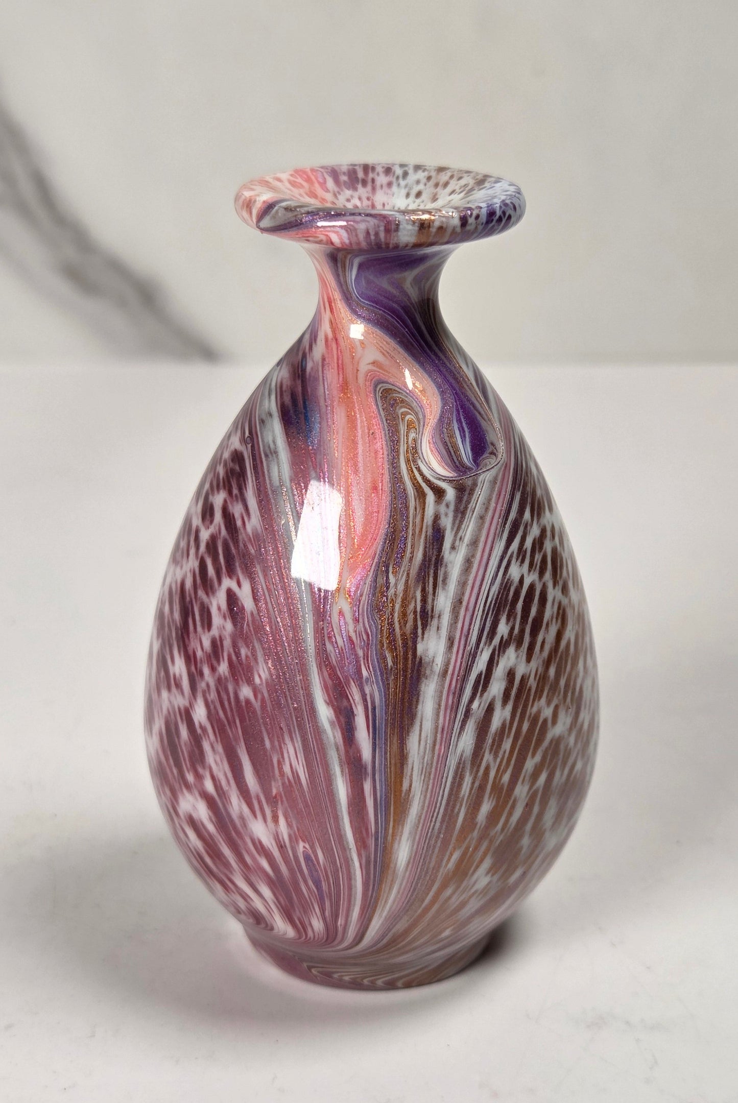 A small decorative glass vase with a rounded bulbous body and a flared rim, featuring a marbled swirl pattern of purple, pink, white, and brown on a light gray marble surface.