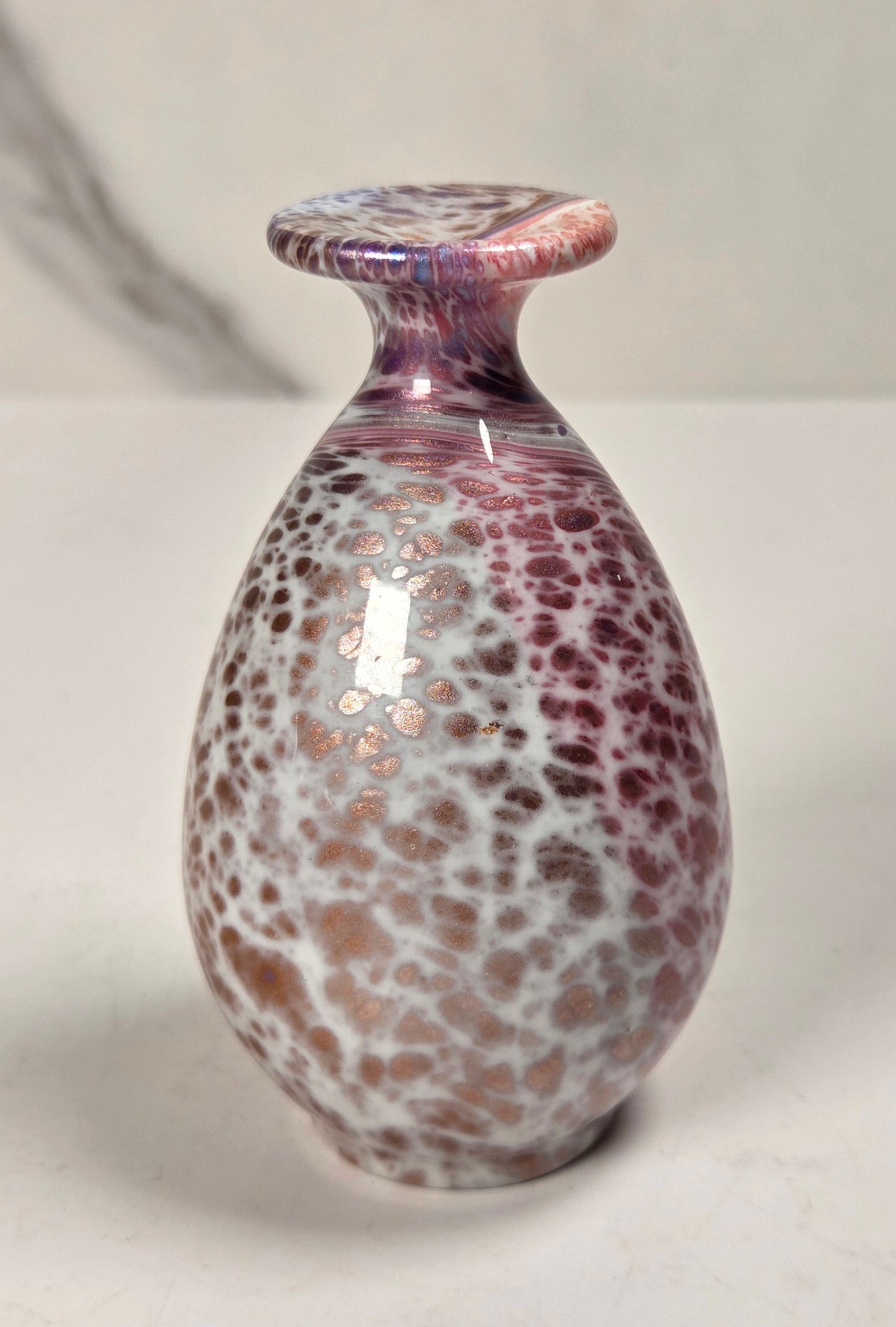 A small decorative glass vase with a rounded bulbous body, narrow neck, and flared top, featuring a mottled pattern of white, pink, and purple with metallic rose-gold flecks, set against a light gray background.