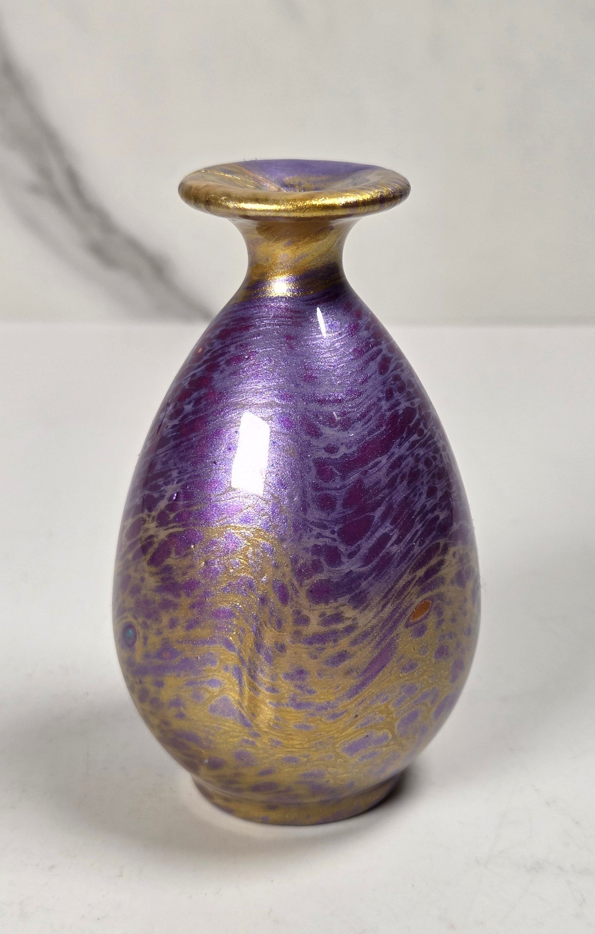 A small decorative ceramic vase with a rounded teardrop shape, featuring a metallic purple and gold marbled pattern, a gold-toned rim, and a matching gold-toned base, set against a light gray marble-patterned background.