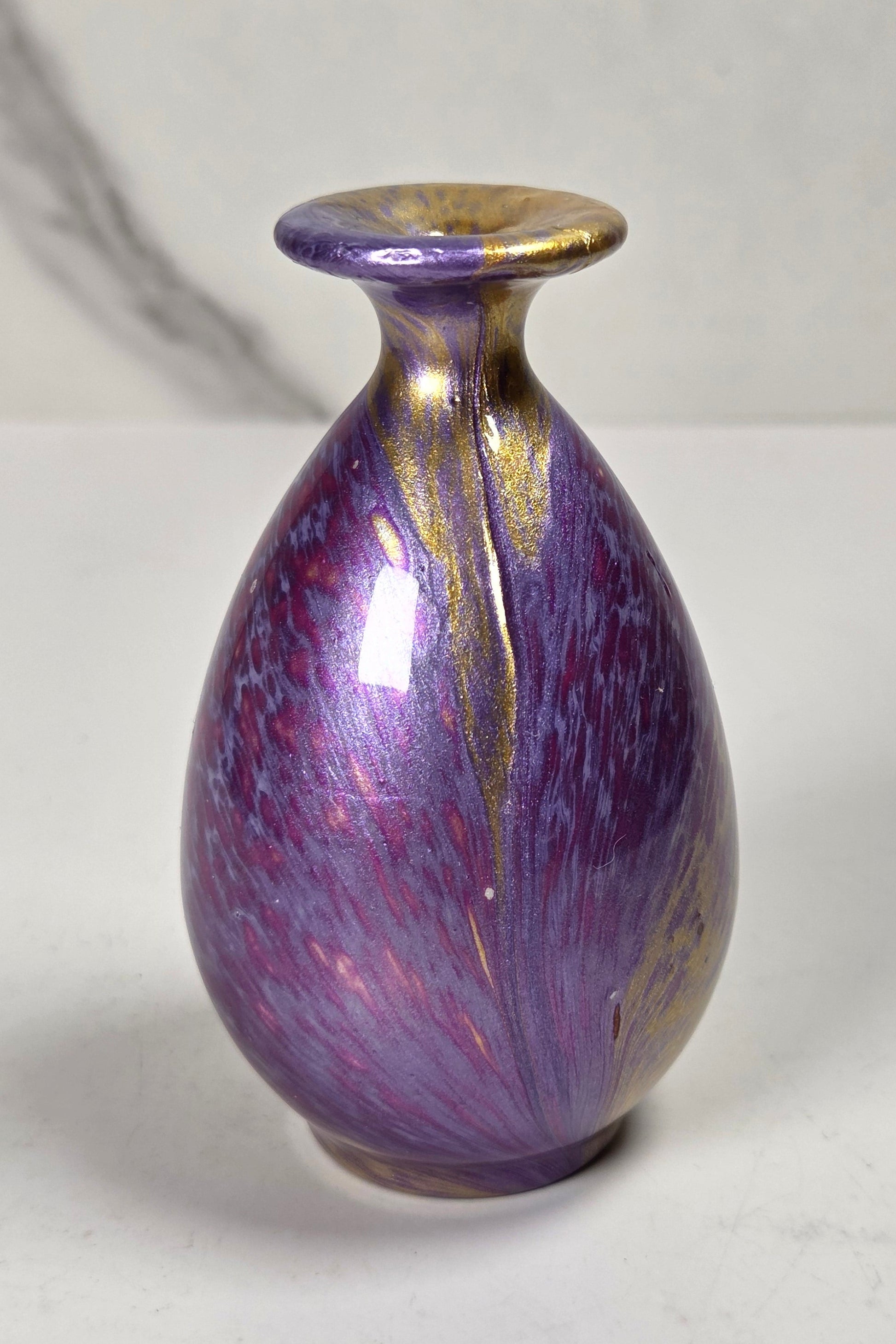 A small ceramic bud vase with a glossy purple and iridescent lavender body, accented with metallic gold leaf details on the rim and sides, set against a light gray marble-patterned background.