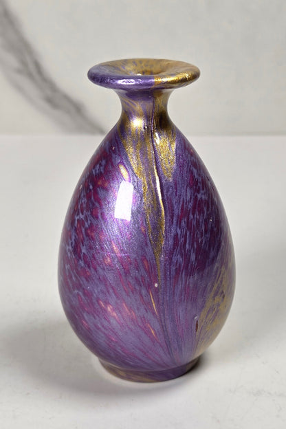 A small ceramic bud vase with a glossy purple and iridescent lavender body, accented with metallic gold leaf details on the rim and sides, set against a light gray marble-patterned background.