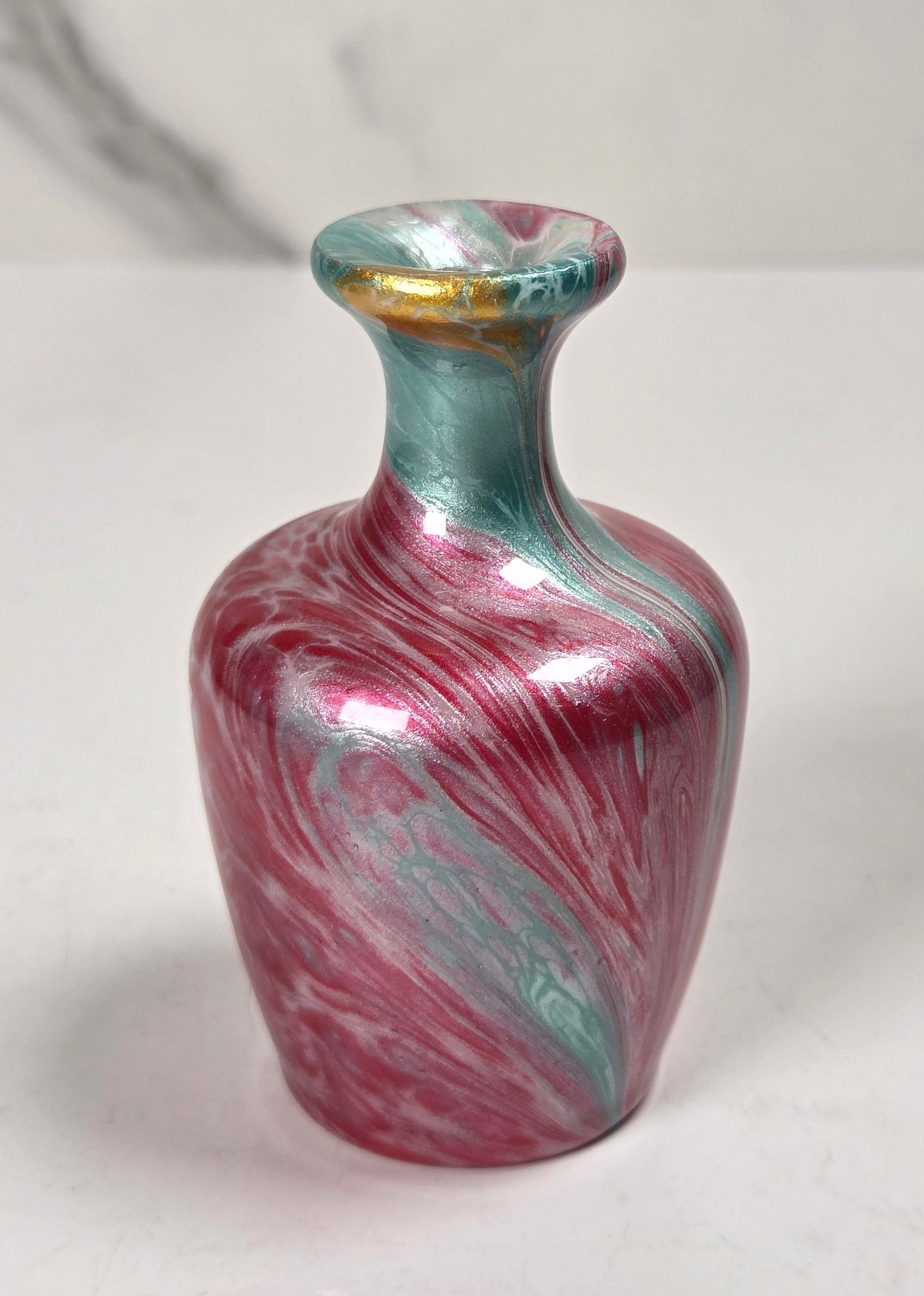 A small decorative vase with a marbled swirl pattern in metallic pink, teal, and silver, featuring a gold accent on the rim, set against a light gray marble-patterned background.