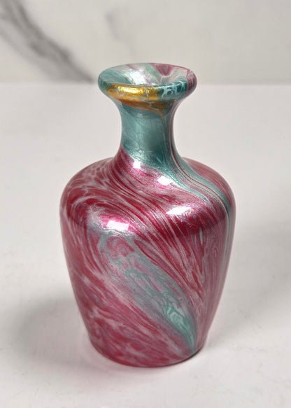 A small decorative vase with a marbled swirl pattern in metallic pink, teal, and silver, featuring a gold accent on the rim, set against a light gray marble-patterned background.