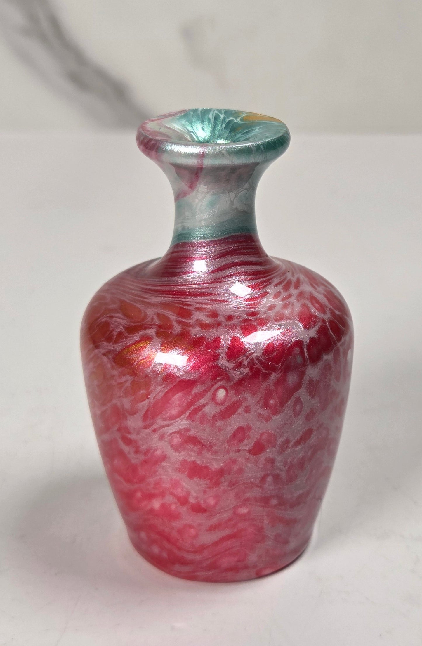 A small decorative glass vase with a rounded body, narrow neck, and flared rim, featuring a marbled pink-red and white pattern with a teal-green top section, set against a light gray marble-patterned background.