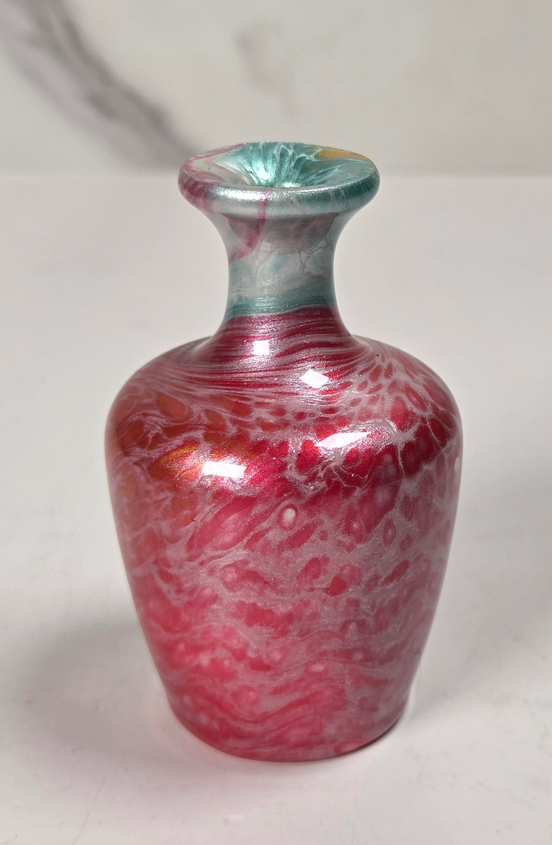 A small decorative glass vase with a rounded body, narrow neck, and flared rim, featuring a marbled pink-red and white pattern with a teal-green top section, set against a light gray marble-patterned background.
