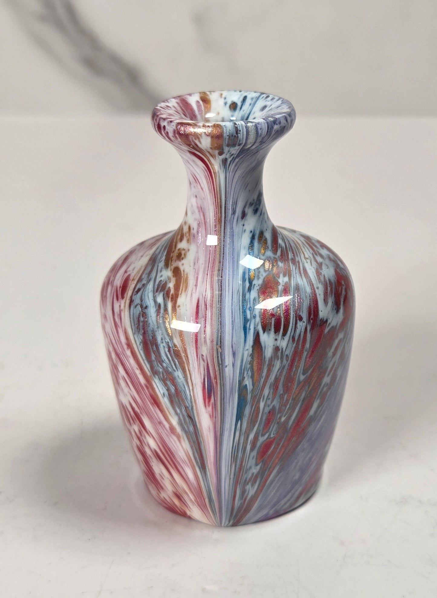 A small decorative vase with a flared neck and rounded body, featuring a marbled swirl pattern in red, blue, white, and hints of gold, set against a light gray marble-patterned background.