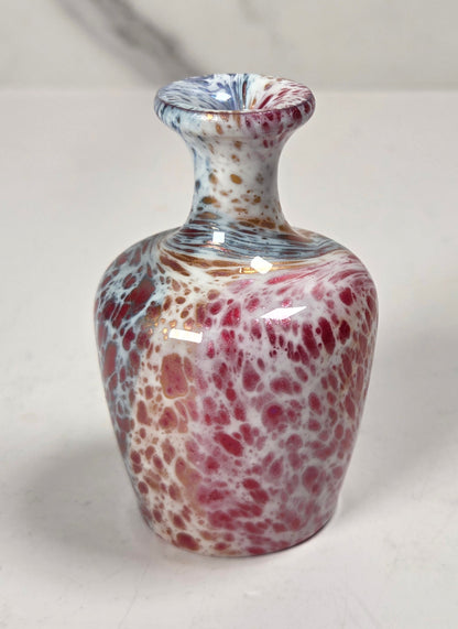 A small hand-blown glass vase with a bulbous body, featuring a marbled pattern of red, pink, white, and blue glass, set against a light gray marble background.