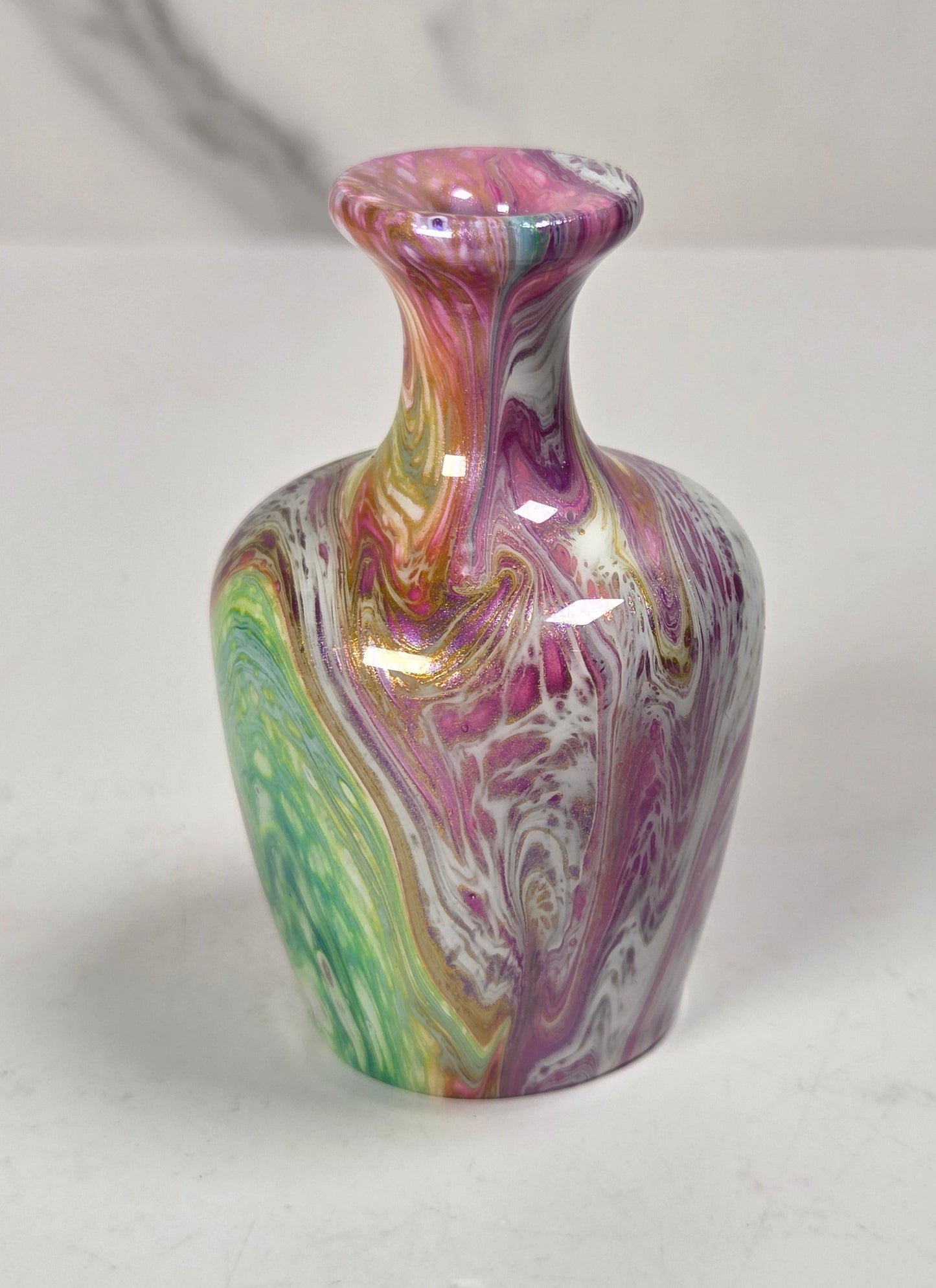 A small glossy ceramic vase with a marbled swirl pattern in pink, purple, green, yellow, and white, set on a light gray surface against a light gray background.