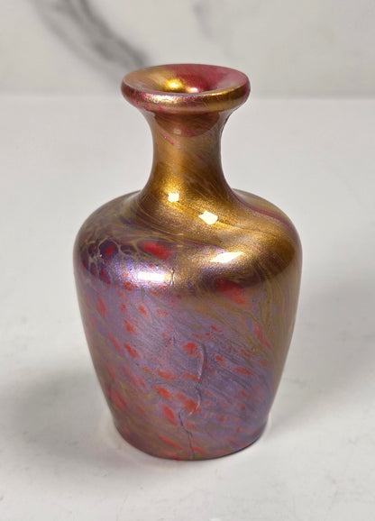 A small iridescent glass vase with a metallic gold and purple body, featuring red marbling and a pinkish-red rim, set against a light gray background.