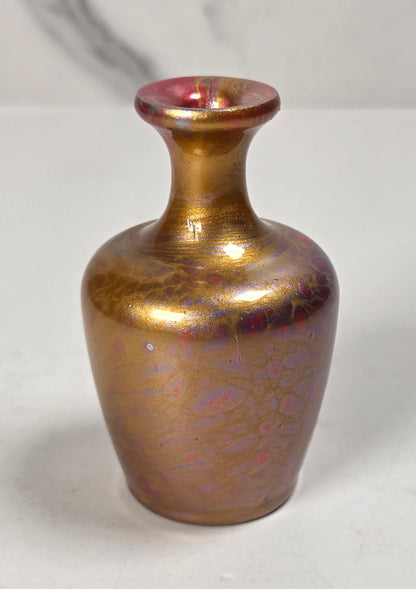 A small iridescent glass vase with a bulbous body and narrow neck, featuring a metallic gold finish with pink and purple shifts, set against a light gray marble-patterned background.