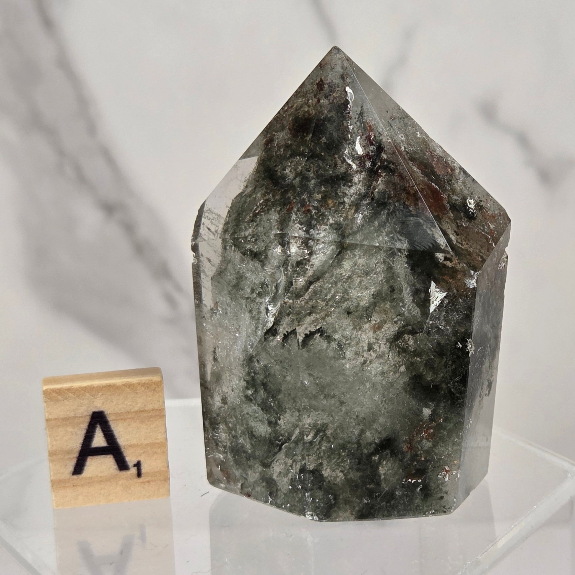 A natural pointed crystal mineral specimen with dark gray and black internal inclusions, displayed on a clear acrylic stand next to a small wooden letter tile showing 'A1' against a white marble background.