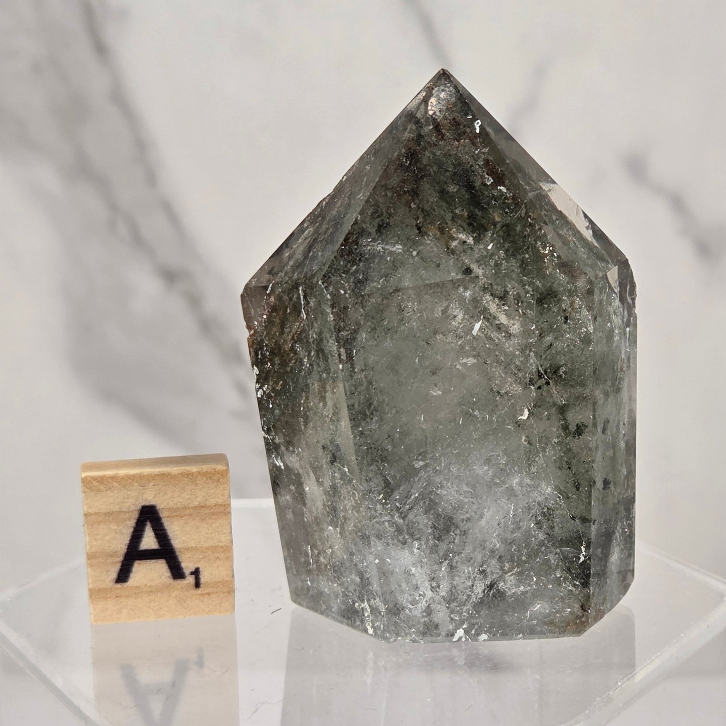 A natural pointed crystal mineral specimen with a smoky gray and translucent quality, displayed on a clear acrylic stand next to a wooden Scrabble tile with the letter 'A'.