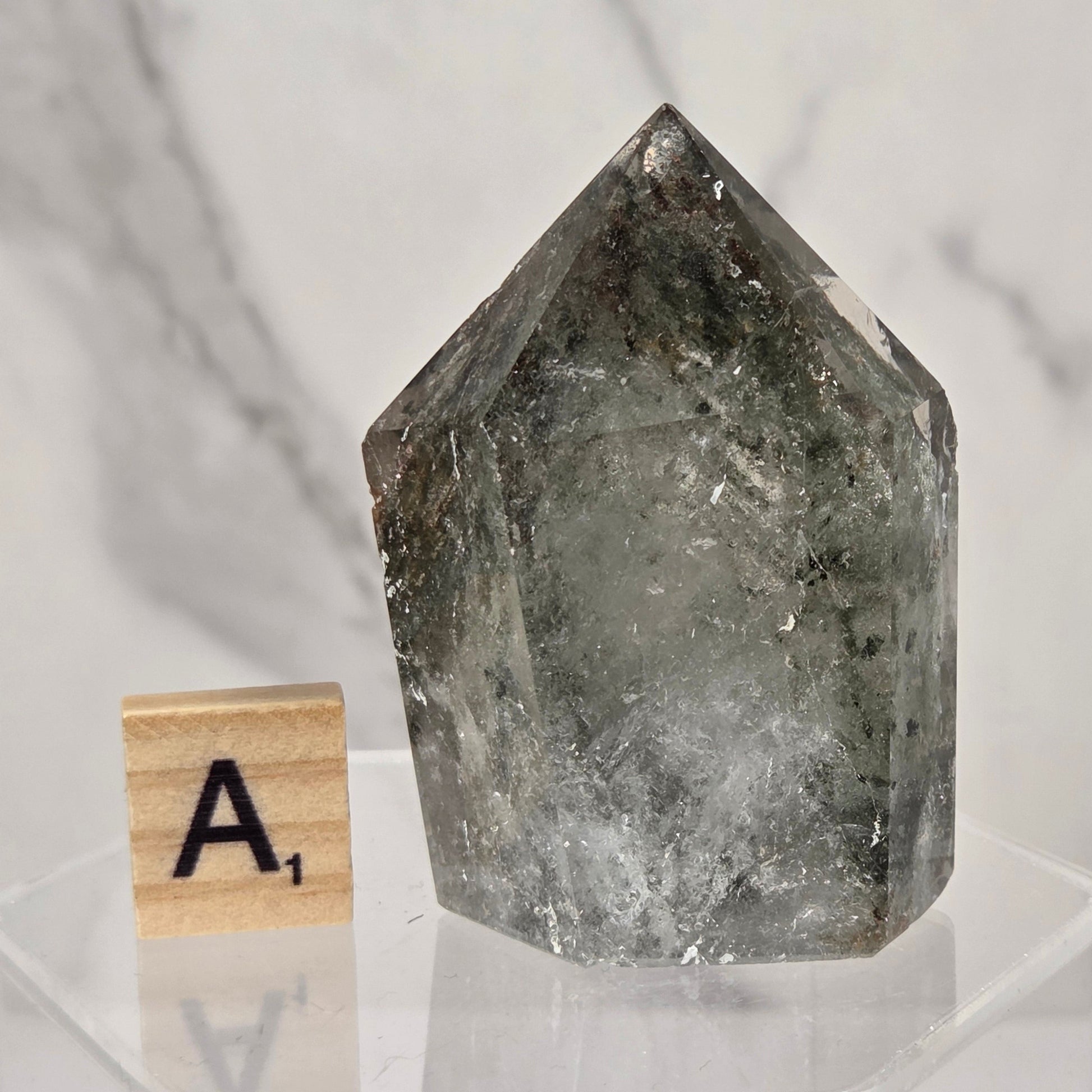 A natural pointed crystal mineral specimen with a smoky gray and translucent quality, displayed on a clear acrylic stand next to a wooden Scrabble tile with the letter 'A'.