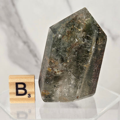 A raw mineral crystal point with a dark gray and green coloration, displayed on a clear acrylic stand next to a wooden Scrabble tile showing the letter 'B'.