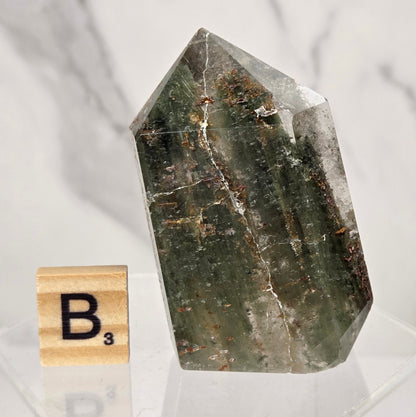 A natural pointed crystal stone with dark green, gray, and brown veining, displayed on a clear acrylic stand next to a wooden Scrabble tile showing the letter B.