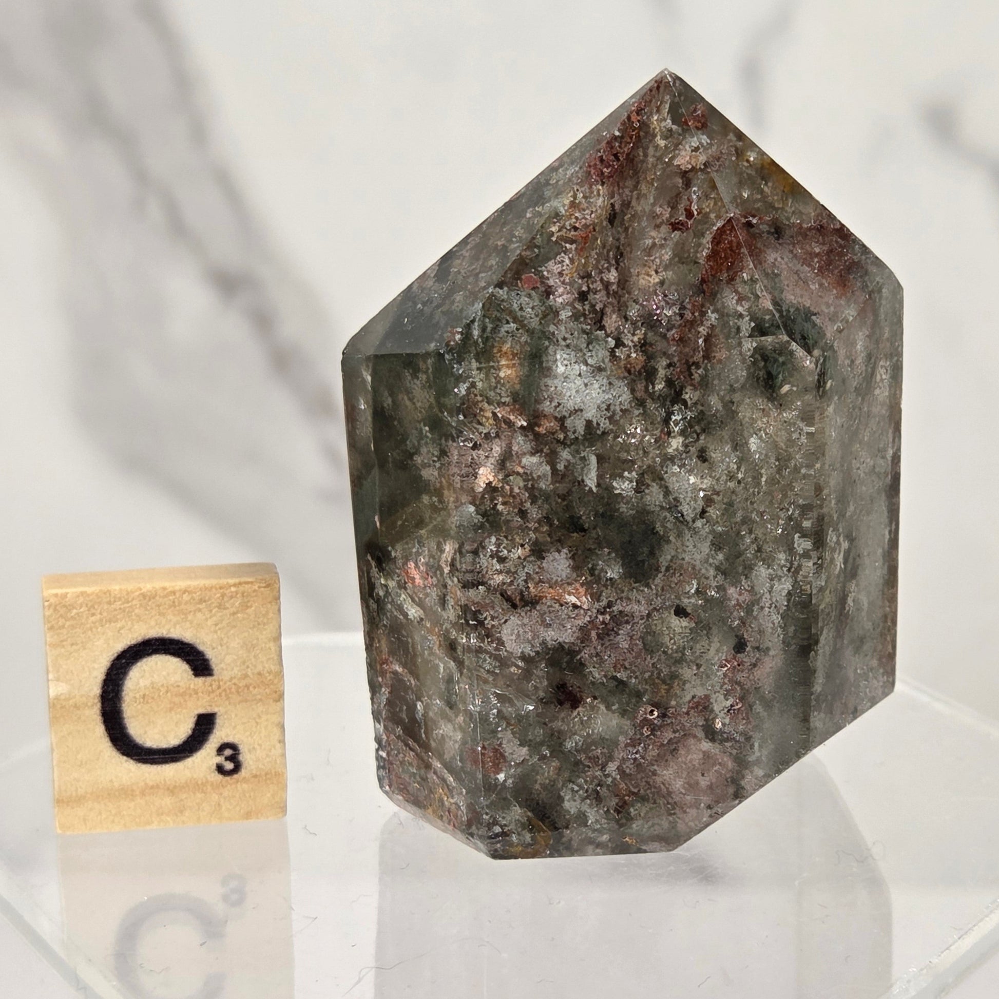 A faceted natural crystal stone with a mottled dark gray, brown, and coppery mineral pattern, displayed on a clear acrylic stand next to a wooden Scrabble tile marked C3, against a light marble background.