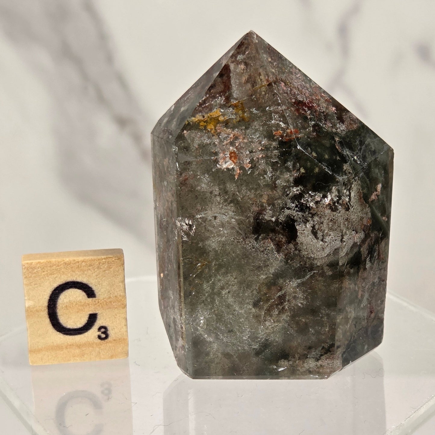 A pointed crystal mineral specimen with a mottled dark gray, green, and brown texture, displayed on a clear acrylic stand next to a wooden Scrabble tile showing the letter C3.
