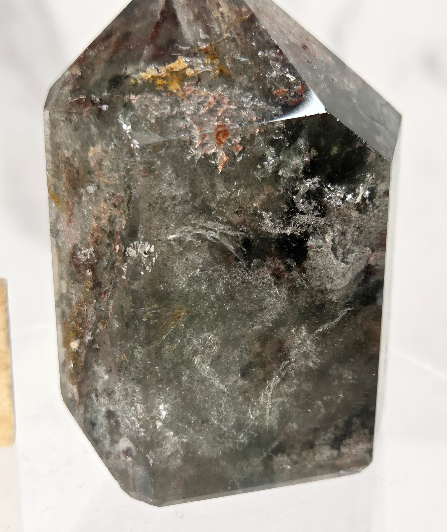 A faceted crystal point, likely smoky quartz, featuring a dark gray to black body with prominent white and gray crystalline inclusions and subtle brownish-orange mineral flecks, displayed on a white surface.