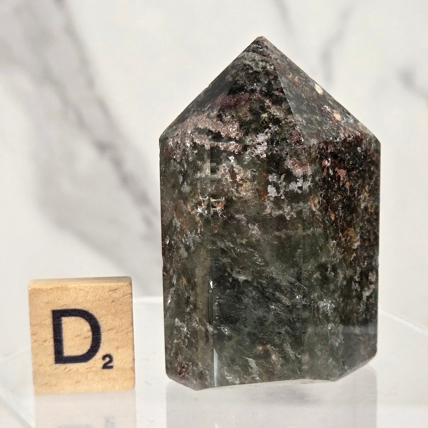 A dark, polished natural crystal tower with a pointed top, displayed on a clear acrylic stand next to a small wooden Scrabble tile showing the letter 'D'.