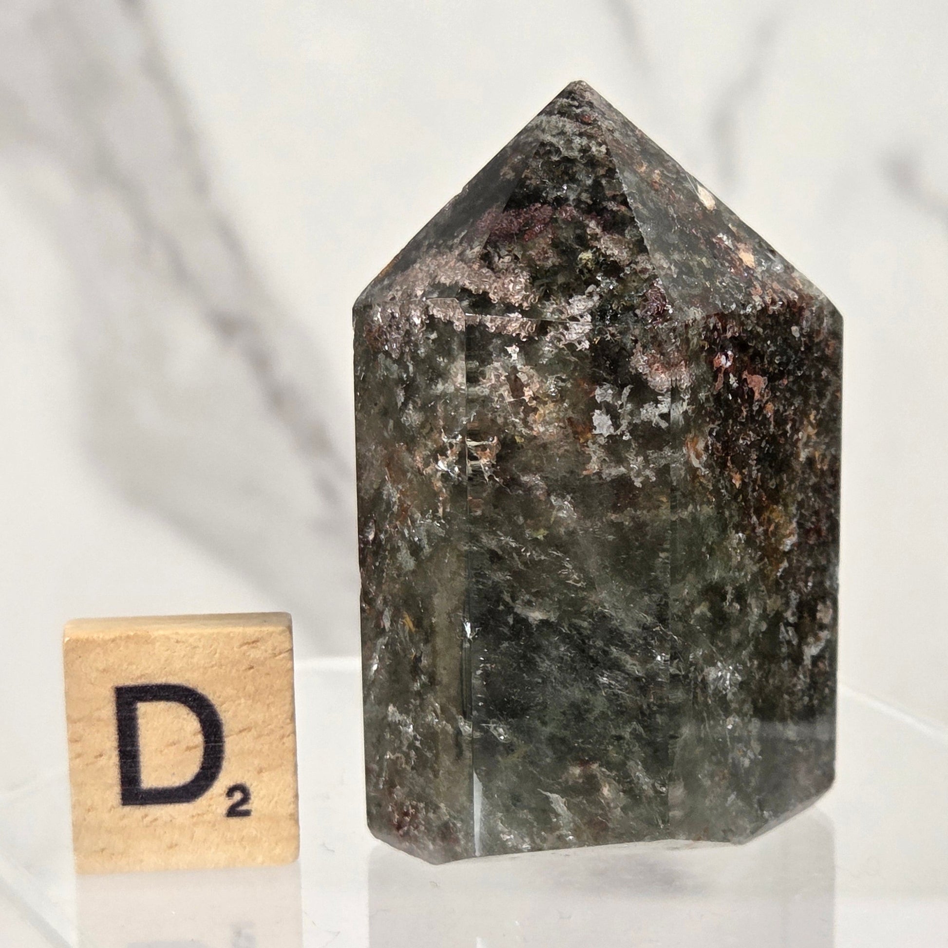 A dark, polished natural crystal tower with a pointed top, displayed on a clear acrylic stand next to a small wooden Scrabble tile showing the letter 'D'.