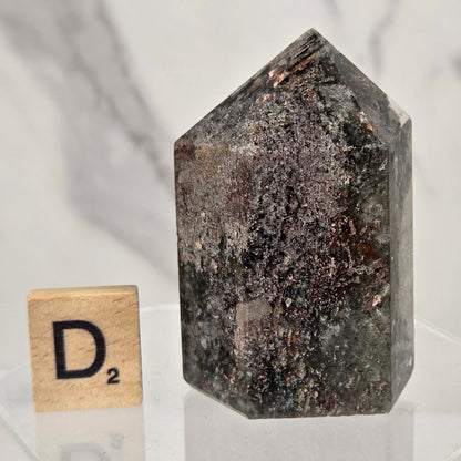 A dark natural crystal point with a rough, speckled texture, displayed on a clear acrylic stand next to a wooden Scrabble tile showing the letter 'D' and the score 'D2'.