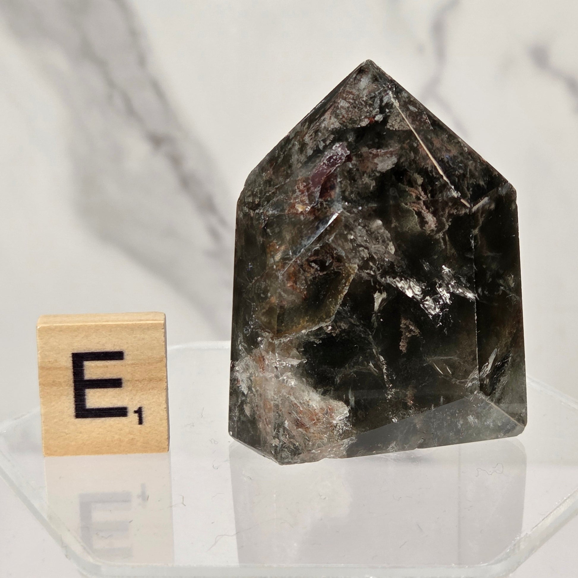 A dark smoky quartz crystal tower with natural inclusions, displayed on a clear acrylic stand next to a wooden Scrabble tile showing the letter 'E' with a score of 1, against a white marble background.