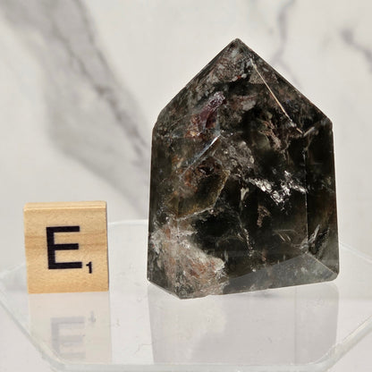 A dark smoky quartz crystal tower with natural inclusions, displayed on a clear acrylic stand next to a wooden Scrabble tile showing the letter 'E' with a score of 1, against a white marble background.