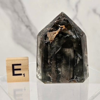 A dark smoky quartz crystal tower with natural inclusions, displayed on a clear acrylic block next to a wooden Scrabble tile showing the letter 'E' and a 1.
