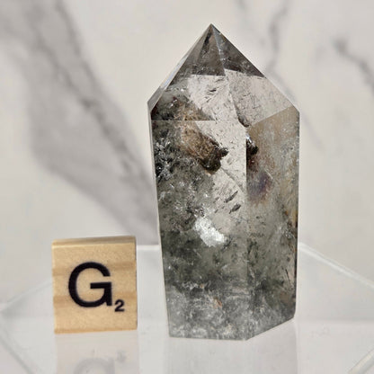 A clear pointed crystal tower with internal inclusions, displayed on a clear acrylic stand next to a wooden Scrabble tile showing the letter G and the score 2, against a white marble-patterned background.