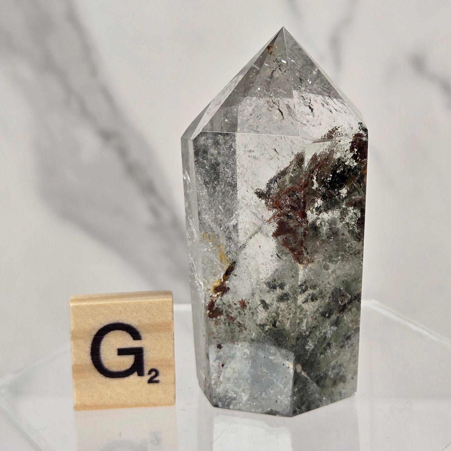 A clear pointed crystal tower with gray and dark brown inclusions, displayed on a clear acrylic stand next to a wooden Scrabble tile marked G2, against a light gray marble-patterned background.