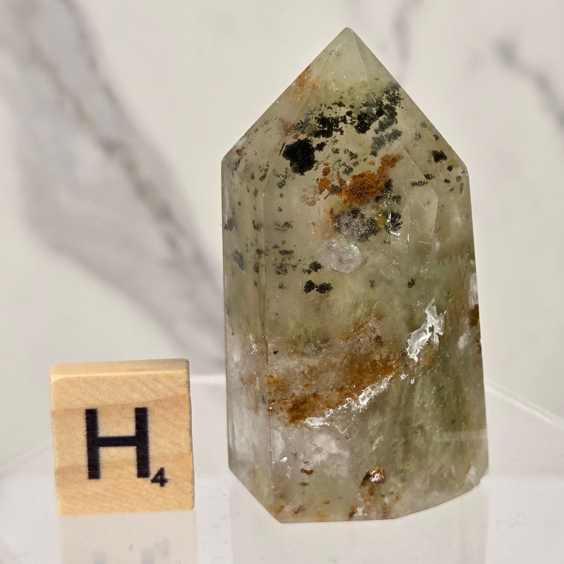 A natural pointed crystal stone with greenish-gray translucent body and brown, black, and white inclusions, displayed next to a wooden Scrabble tile showing the letter H on a clear acrylic stand.