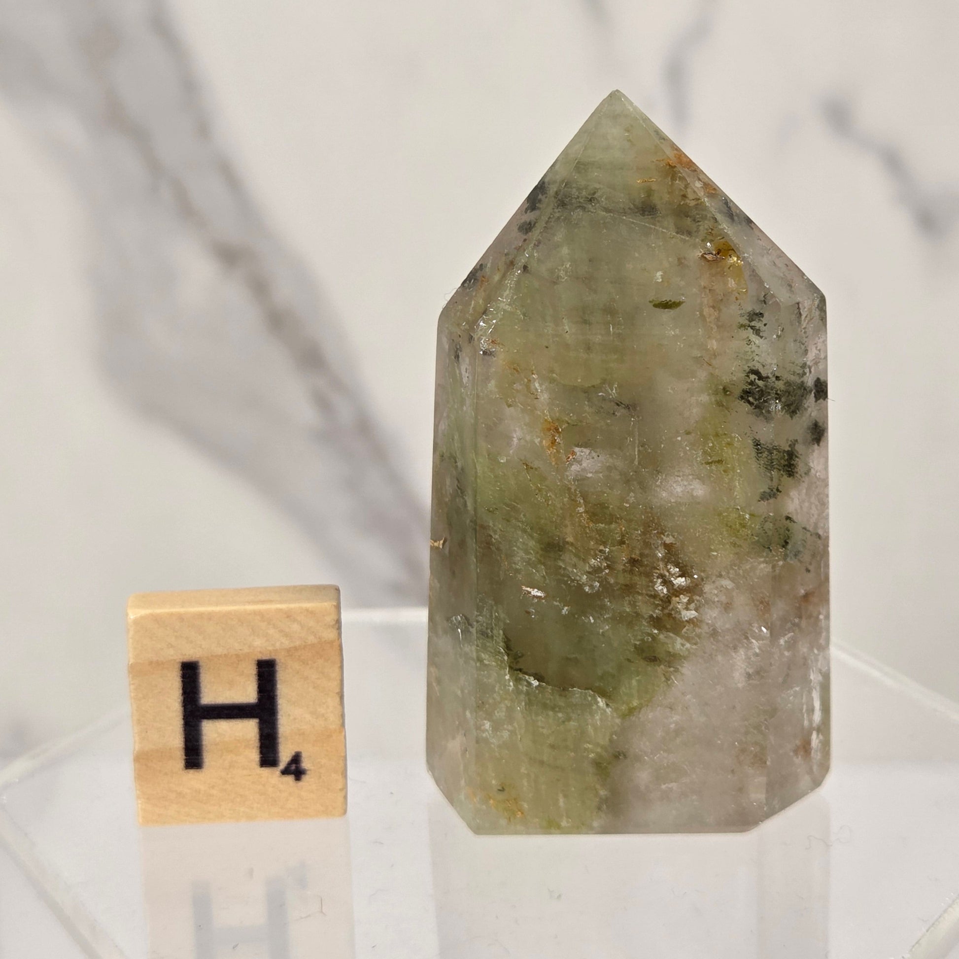 A pointed green crystal tower with natural inclusions, displayed next to a wooden Scrabble tile showing the letter H on a clear acrylic stand against a marble background.