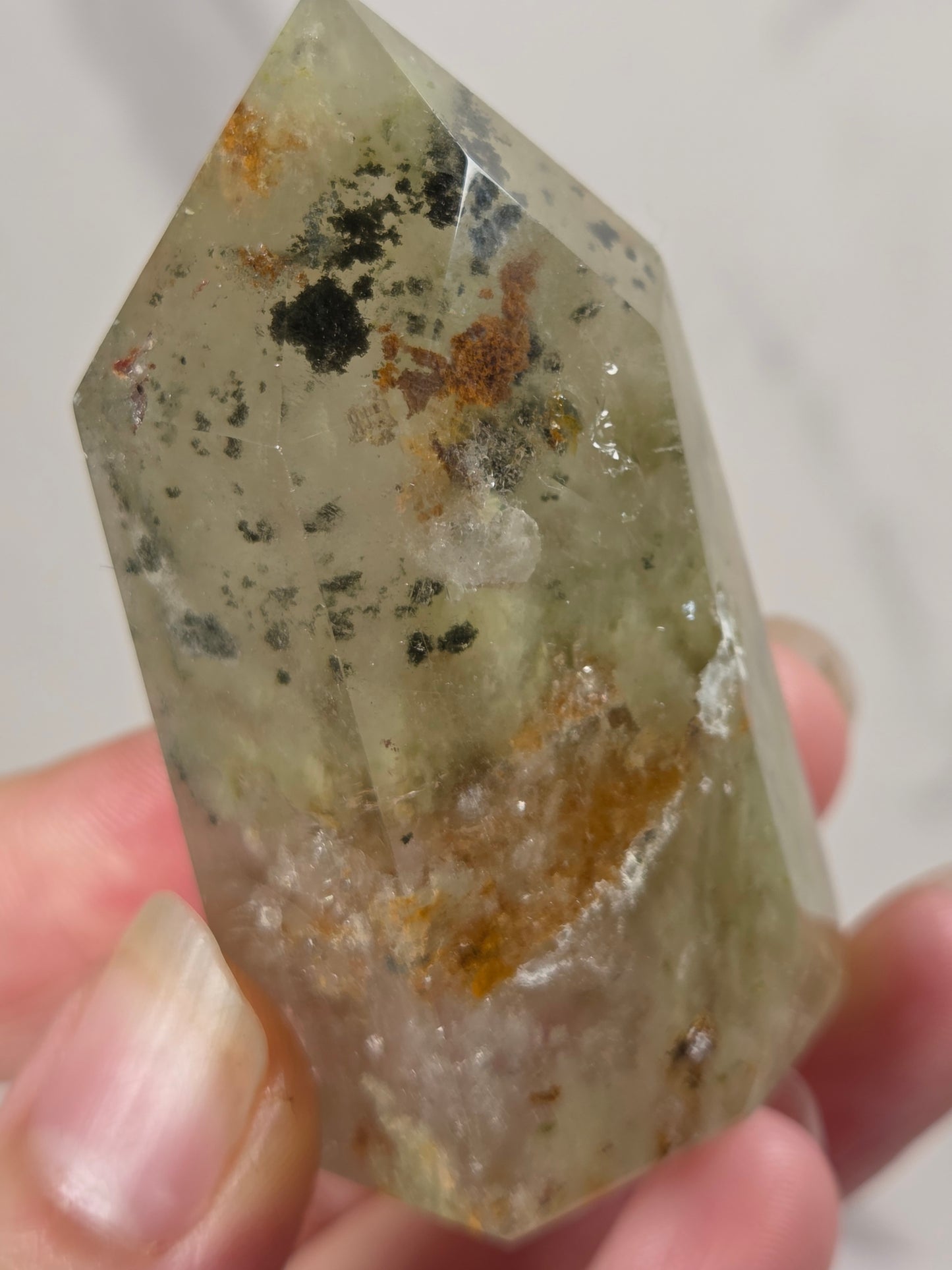 A hand holding a natural quartz point crystal with greenish-yellow tones, dark inclusions, and orange-brown mineral deposits, shown against a light marble background.