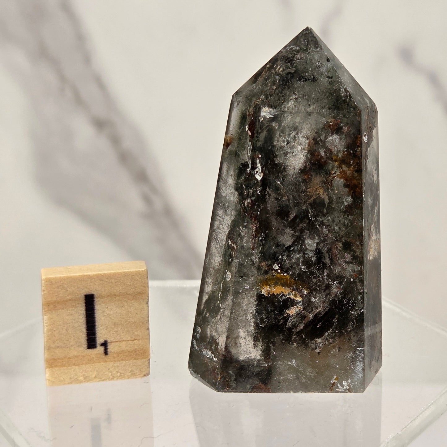 A polished natural crystal point, likely quartz, with dark gray, black, and brown inclusions, displayed on a clear acrylic stand next to a small wooden block with the number 1.