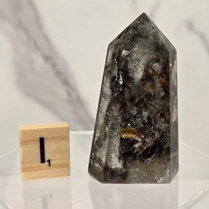 A polished natural crystal point, likely quartz, with dark gray, black, and brown inclusions, displayed on a clear acrylic stand next to a small wooden block with the number 1.