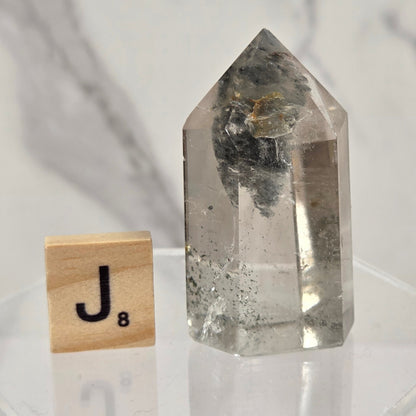 A clear pointed crystal tower with internal dark inclusions, displayed on a clear acrylic base next to a wooden Scrabble tile showing the letter J.