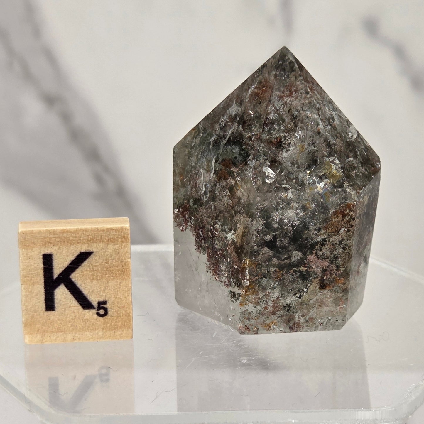 A natural pointed crystal gemstone with a mottled gray, brown, and white speckled pattern, displayed on a clear acrylic stand next to a wooden Scrabble tile spelling 'K' with a 5.